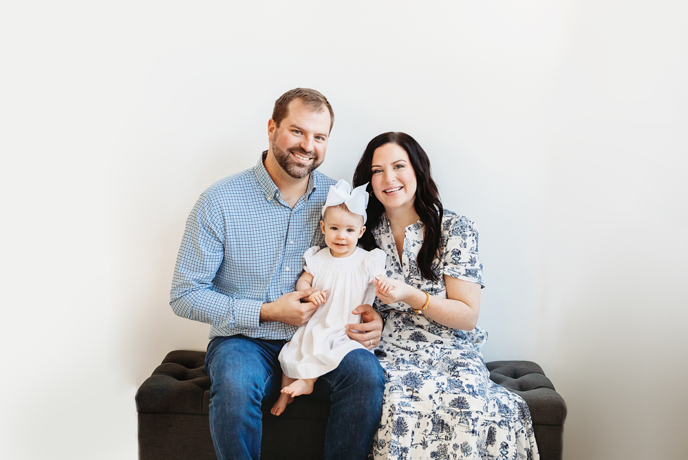 Full family portrait on dark bench, Elsie in center wearing white dress and bow, bright studio backdrop at Reflection Images by Tracy Rowell in Florence SC