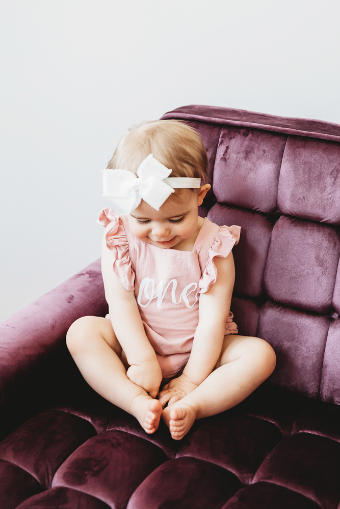 Grace in a pink 'one' romper with ruffled sleeves, sitting cross-legged looking down on a plum velvet chair at Reflection Images by Tracy Rowell in Florence SC
