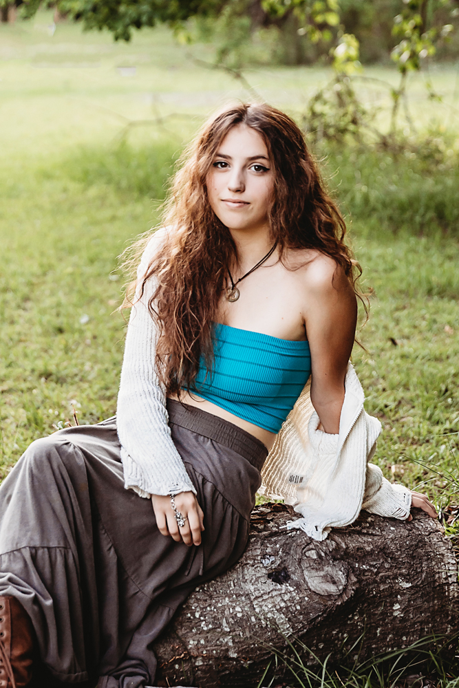 Eve seated casually on a tree log in skirt and sweater, turquoise bandeau top, relaxed expression in grassy clearing at Reflection Images by Tracy Rowell in Florence SC