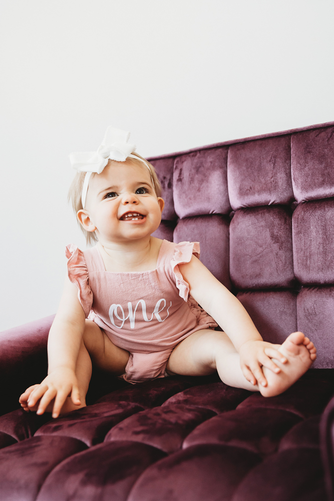 Grace in a 'one' romper smiling brightly with legs stretched out on a plum velvet chair at Reflection Images by Tracy Rowell in Florence SC
