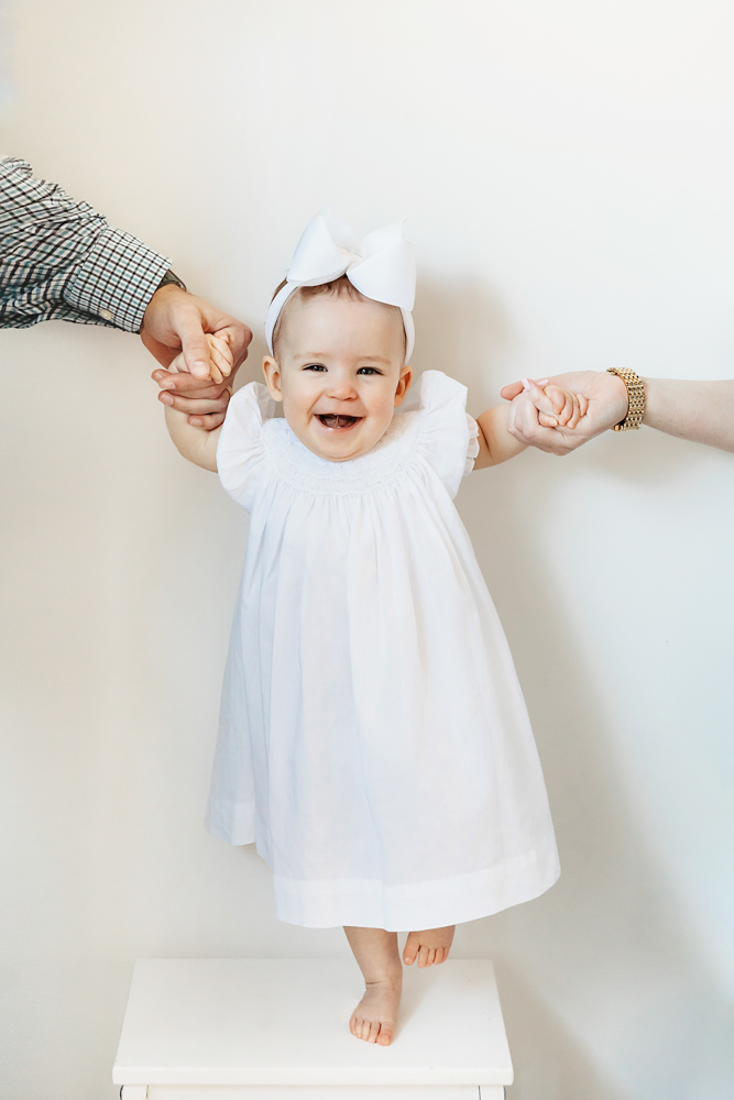 Elsie standing on small stool in white dress and bow, parents’ hands holding hers for balance, milestone photo at Reflection Images by Tracy Rowell in Florence SC