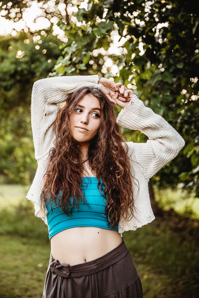 Eve raising arms above head with long wavy hair, turquoise top, cream sweater, forest greenery backdrop at Reflection Images by Tracy Rowell in Florence SC