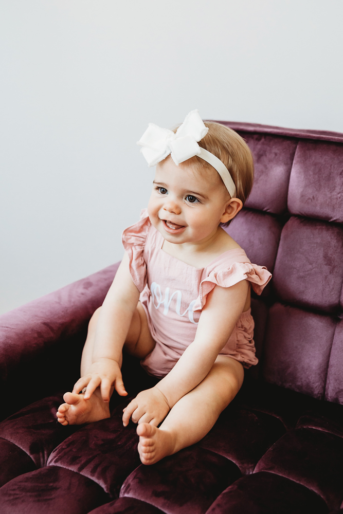Grace smiling off to the side while seated on a velvet purple chair in a pink romper with 'one' text at Reflection Images by Tracy Rowell in Florence SC