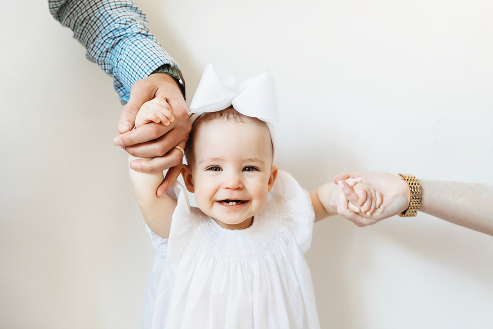 Close-up of Elsie smiling while holding both parents’ hands, white dress and bow, clean studio wall backdrop at Reflection Images by Tracy Rowell in Florence SC