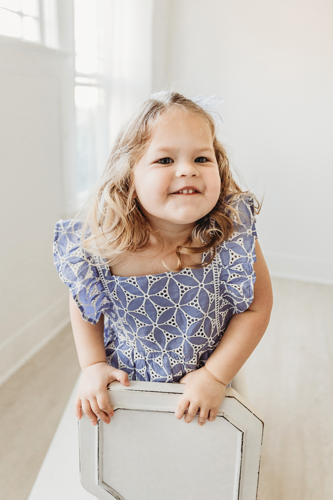 Livi Kates toddler sister leans on a chair and smiles sweetly in her lavender birthday dress.