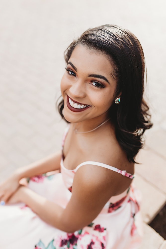 Top-down image of Connie sitting on a brick sidewalk, smiling up in her floral dress with curled hair at Reflection Images by Tracy Rowell in Florence SC