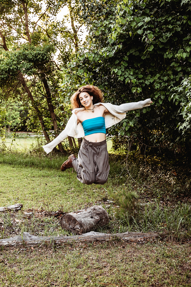 Eve midair jump with arms outstretched, turquoise top, cream sweater, flowing skirt, grassy clearing with trees at Reflection Images by Tracy Rowell in Florence SC