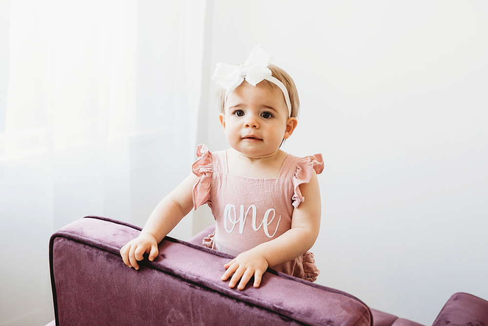 Grace standing beside a plum velvet chair in a pink 'one' romper and white bow near a soft-lit window at Reflection Images by Tracy Rowell in Florence SC