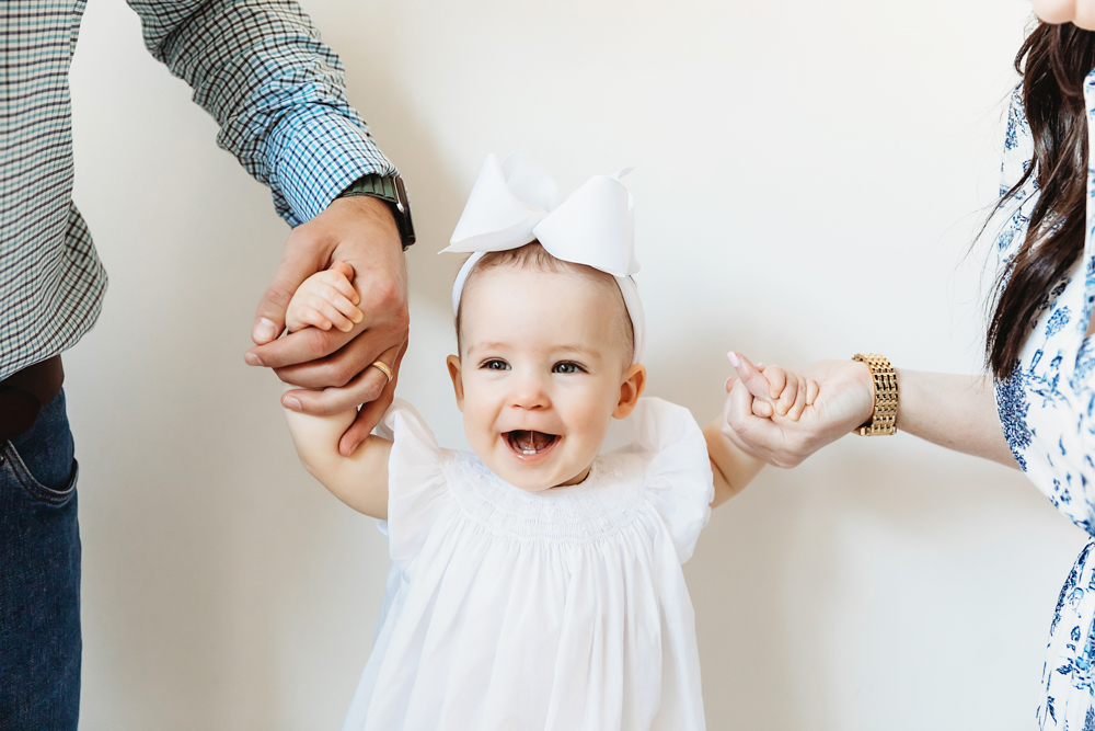 Elsie laughing while holding parents’ hands, white bow and dress, joyful milestone close-up at Reflection Images by Tracy Rowell in Florence SC