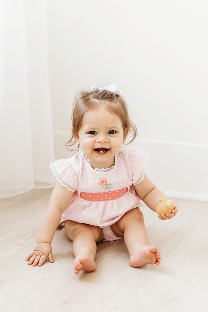 Livi Kate smiles sweetly while sitting on the floor in a pink outfit with a white bow, celebrating her first birthday.