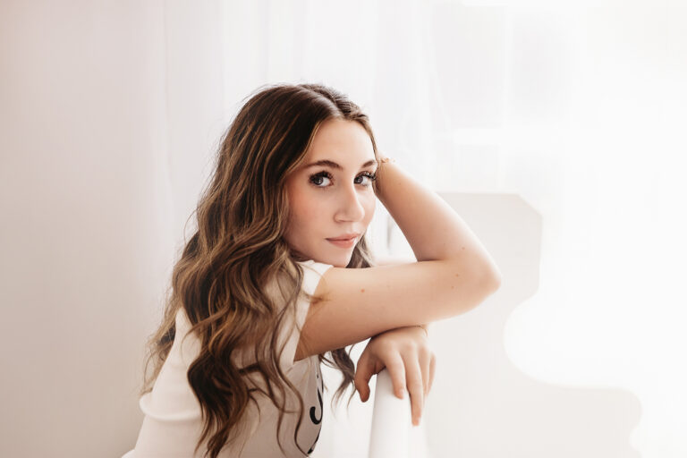 Teen girl leaning thoughtfully against a white stair railing, soft window light illuminating her face and cascading hair.