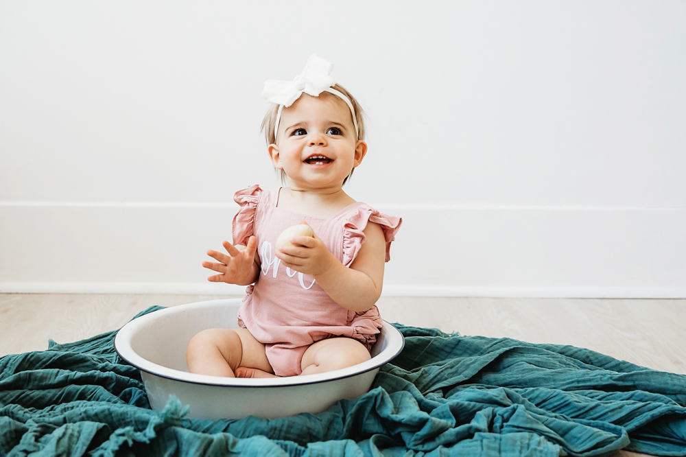 Grace in a 'one' romper sitting in a white enamel bowl over a teal textured blanket, smiling brightly at Reflection Images by Tracy Rowell in Florence SC