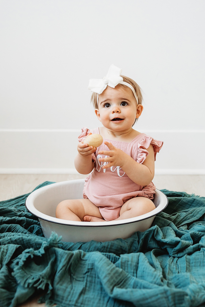 Grace holding a wooden toy while sitting in a bowl in a pink romper and white bow at Reflection Images by Tracy Rowell in Florence SC