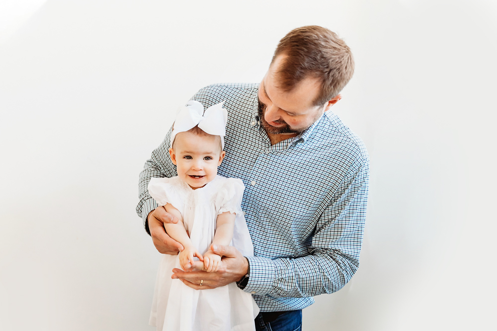 Dad holding Elsie in white dress and bow, both smiling softly, bright studio wall backdrop at Reflection Images by Tracy Rowell in Florence SC