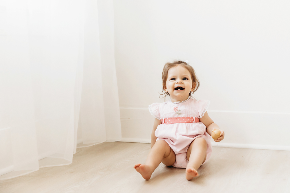 Baby girl wearing a pink dress and smiling while sitting on the floor near a white curtain