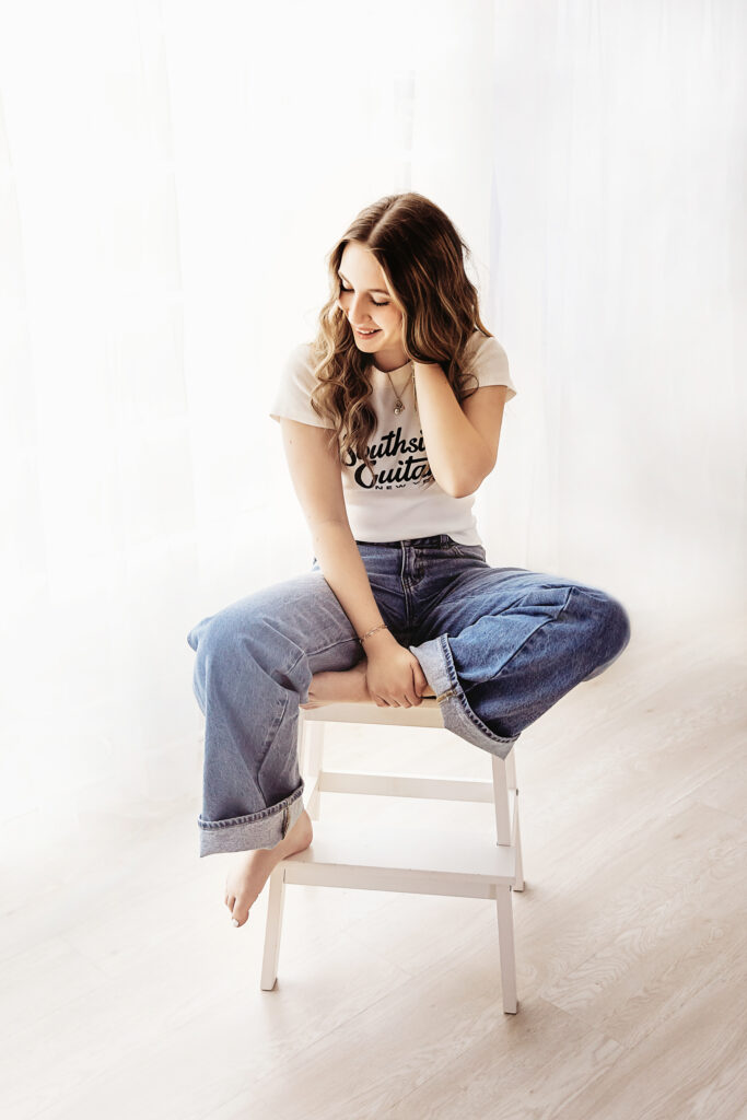 Smiling teen girl sitting cross-legged on a stool in front of soft white curtains, relaxed and joyful expression.