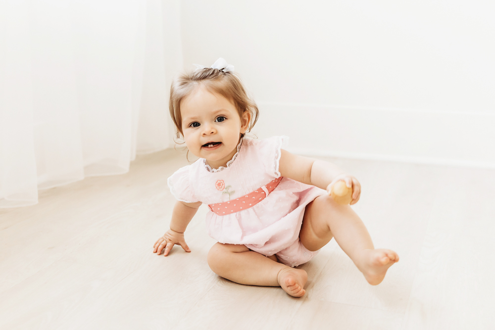 Baby girl crawling on wood floor while wearing a pink birthday dress and holding a small toy