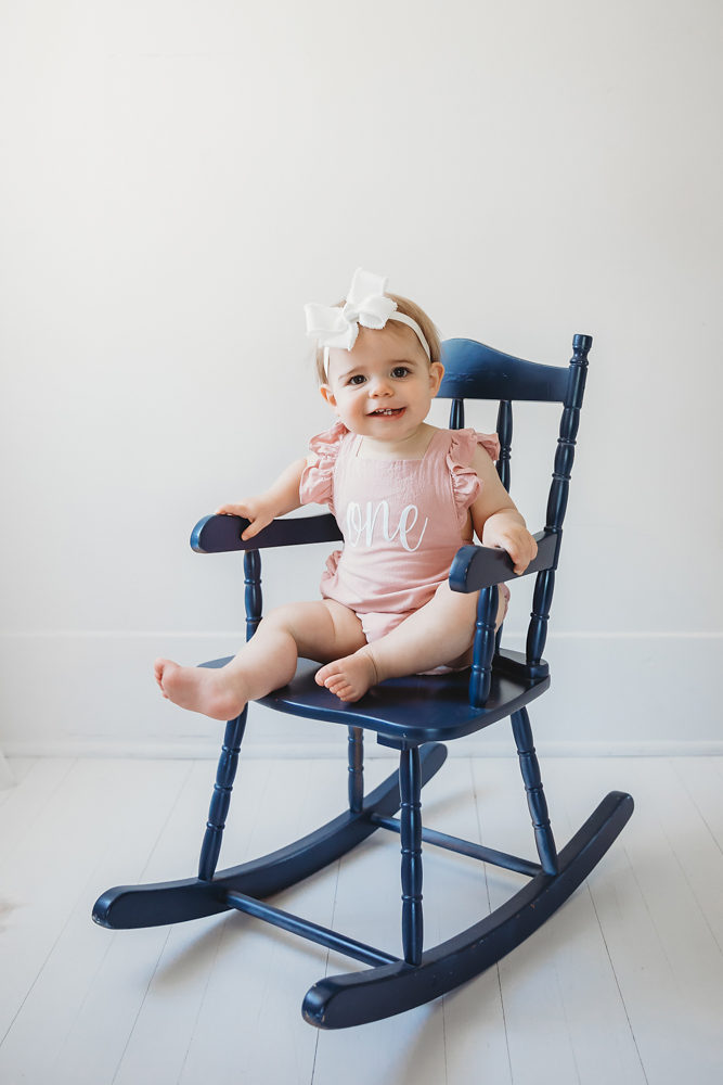 Grace sitting in a navy wooden rocking chair, wearing her pink 'one' romper and smiling with toes out at Reflection Images by Tracy Rowell in Florence SC
