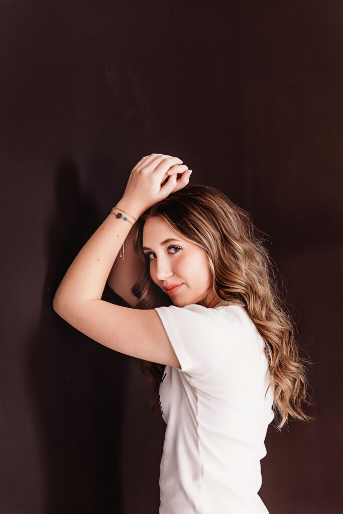 Teen girl leaning back against a black wall with a relaxed smile, soft lighting highlighting her wavy hair.