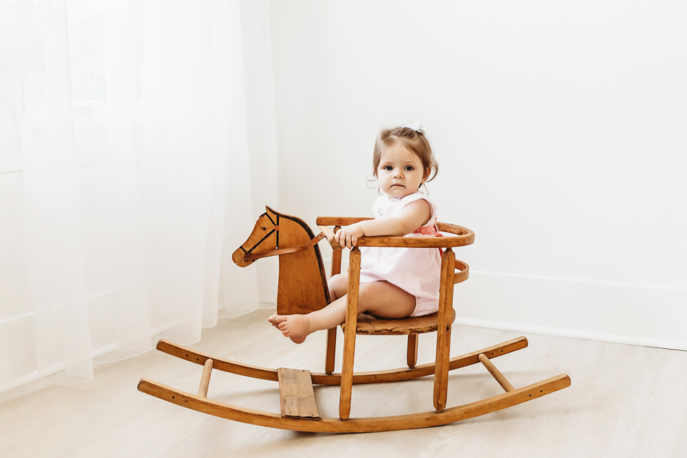 Toddler girl in a pink dress sitting on a vintage wooden rocking horse during birthday shoot