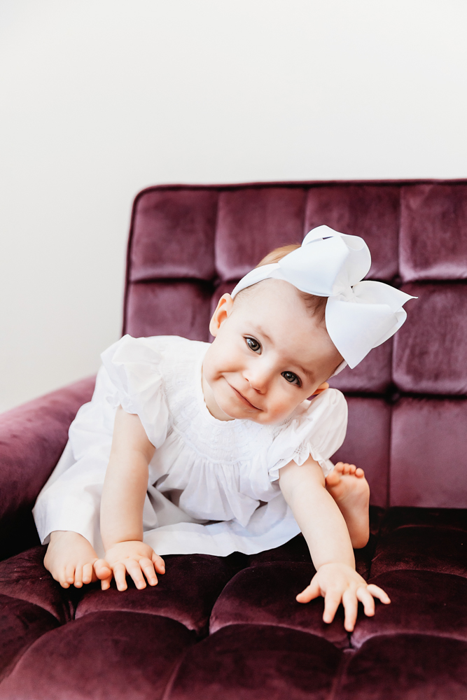 Elsie sitting on plush purple chair, tilting her head playfully, wearing white dress and bow at Reflection Images by Tracy Rowell in Florence SC