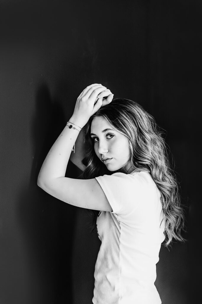 Black and white portrait of a teen girl in profile leaning against a dark wall, wearing a white t-shirt, looking softly toward the camera.
