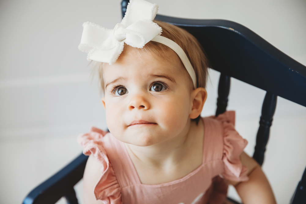 Close-up of Graces curious face in a pink romper and large white bow, seated in a navy rocking chair at Reflection Images by Tracy Rowell in Florence SC
