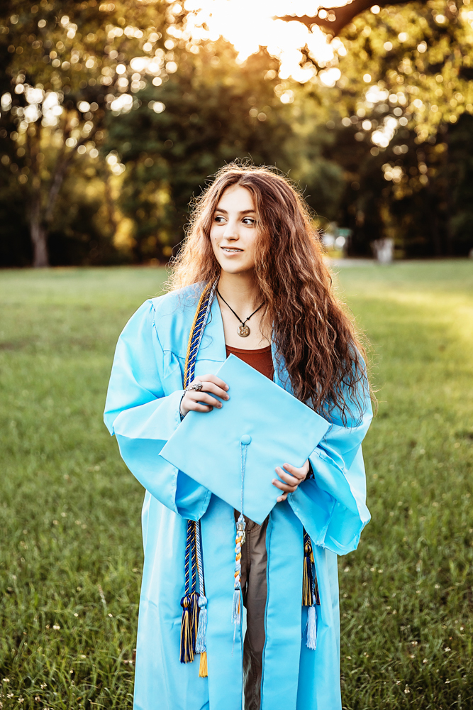 Eve wearing light blue graduation gown holding cap in hands, golden sunset light in grassy field at Reflection Images by Tracy Rowell in Florence SC