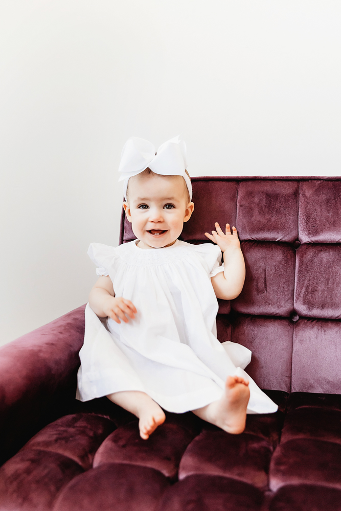 Portrait of Elsie sitting on purple chair, smiling sweetly in white dress and bow at Reflection Images by Tracy Rowell in Florence SC