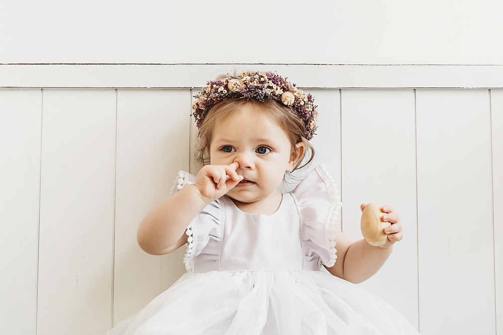 Funny moment of baby girl picking her nose while holding a toy in her white dress and flower crown