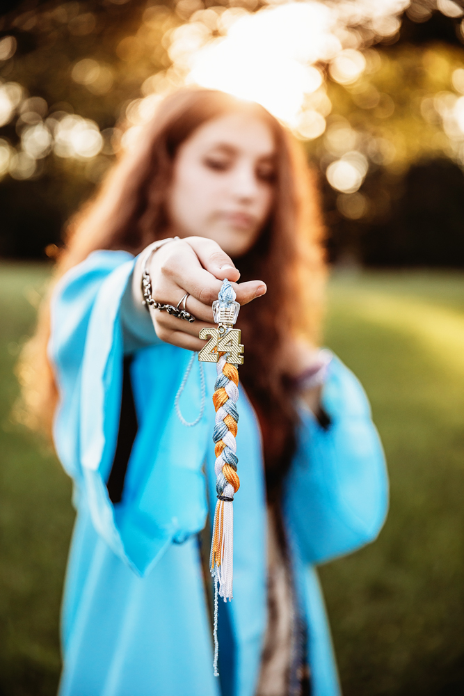 Eve holding 2024 graduation tassel forward in focus, blurred face behind in blue gown with sunlit background at Reflection Images by Tracy Rowell in Florence SC