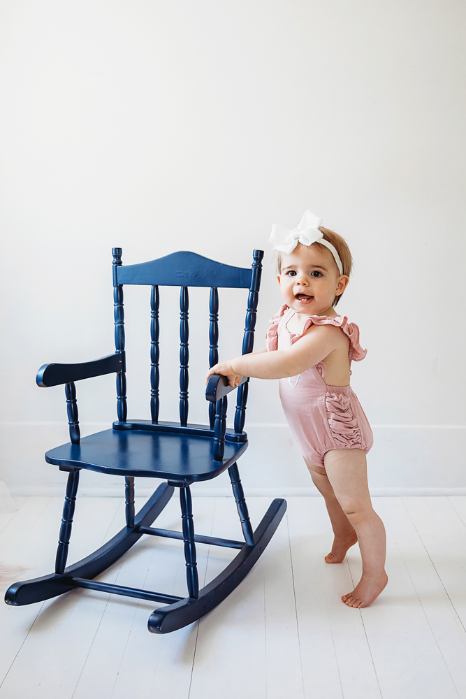 Grace standing barefoot beside a navy rocking chair, one hand on the armrest, wearing a pink romper at Reflection Images by Tracy Rowell in Florence SC