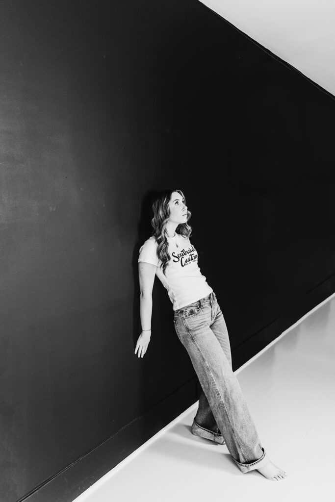 Moody black and white image of teen girl standing barefoot against a black wall, looking upward pensively.
