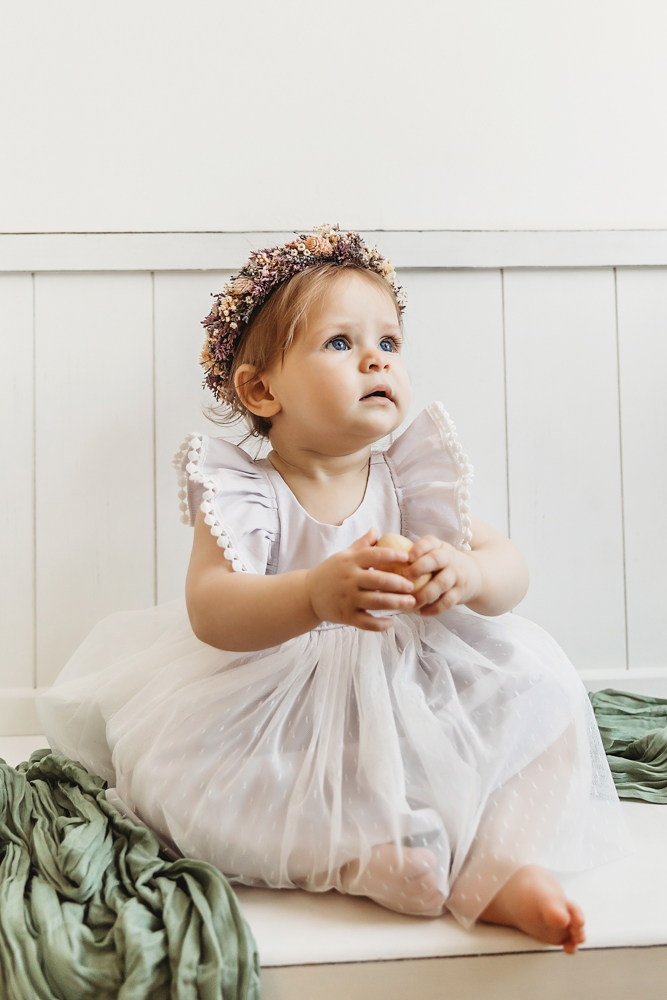 Baby girl looking up while sitting in a white tulle dress and flower crown on a green wrap