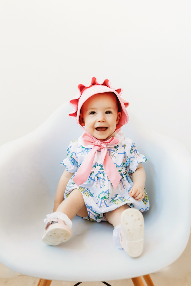 Elsie wearing pink bonnet with large bow, floral romper, sitting in modern white chair at Reflection Images by Tracy Rowell in Florence SC