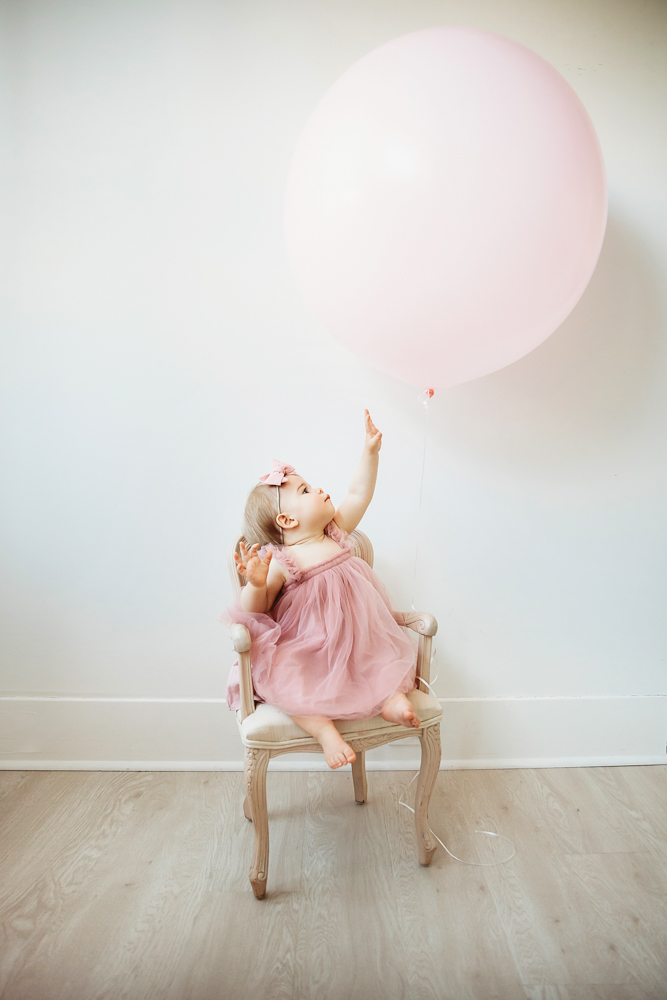 Grace in a pink tulle dress and bow reaching for a large pale pink balloon while sitting on a vintage chair at Reflection Images by Tracy Rowell in Florence SC