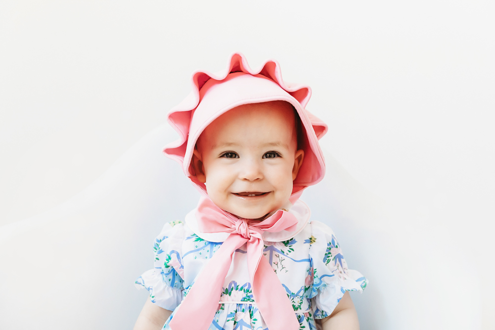 Close-up of Elsie in pink bonnet with ribbon bow, floral romper, smiling against white backdrop at Reflection Images by Tracy Rowell in Florence SC