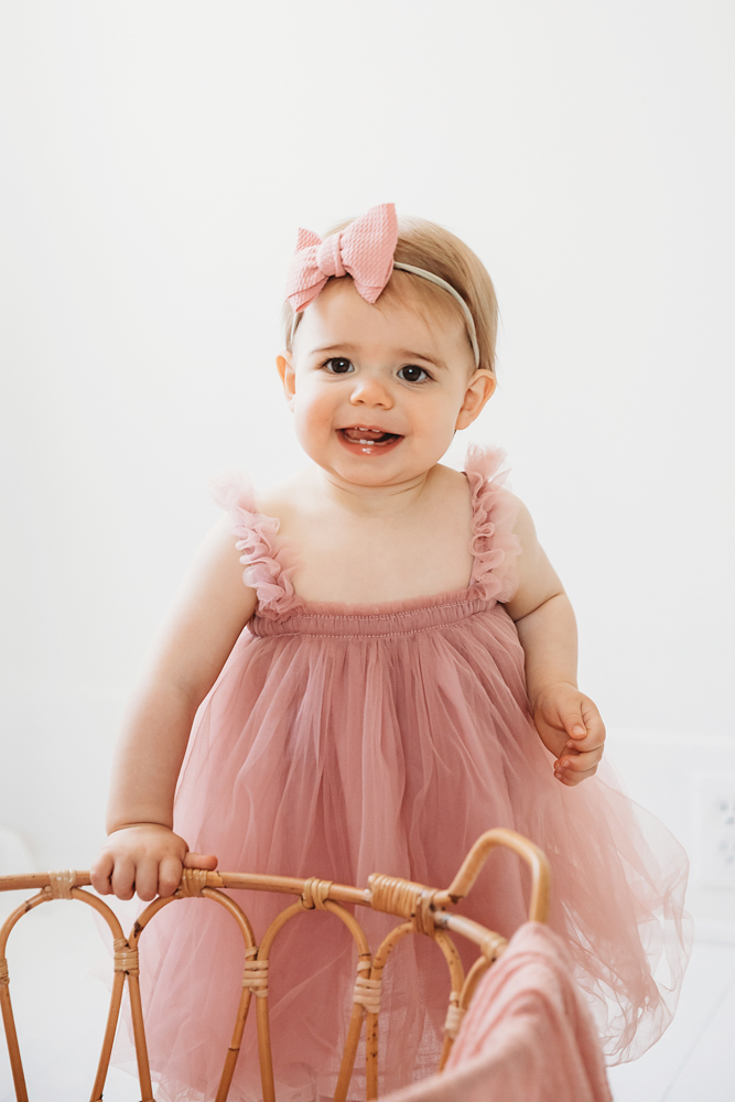 Grace standing by a rattan basket in a pink tulle birthday dress and pink bow, smiling brightly at Reflection Images by Tracy Rowell in Florence SC