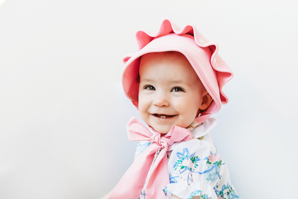 Side portrait of Elsie in floral romper with pink bonnet and ribbon tie, smiling playfully at Reflection Images by Tracy Rowell in Florence SC