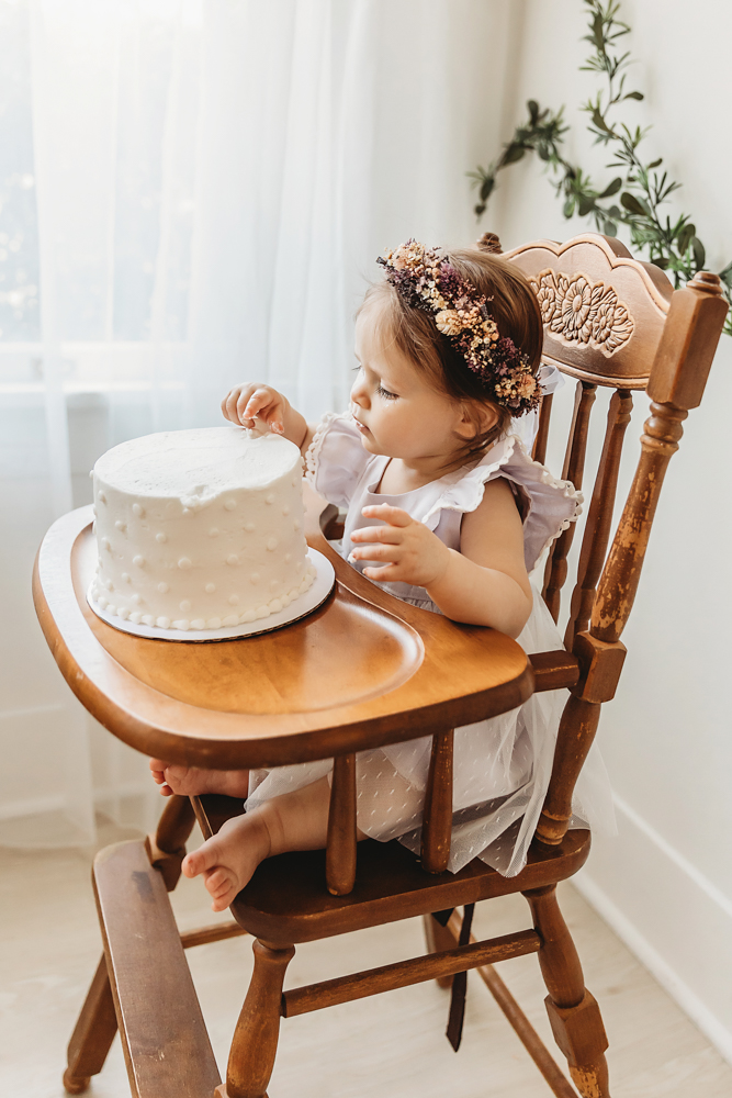 Baby girl in a flower crown and white dress touching her birthday cake while sitting in a vintage high chair