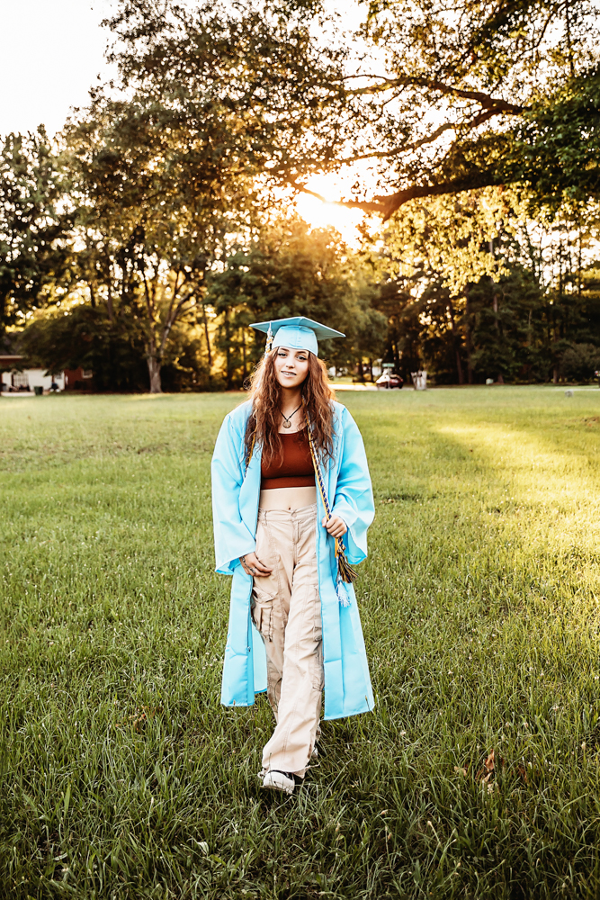 Eve walking in grassy field wearing light blue gown over rust crop top and khaki pants, tassels draped at Reflection Images by Tracy Rowell in Florence SC
