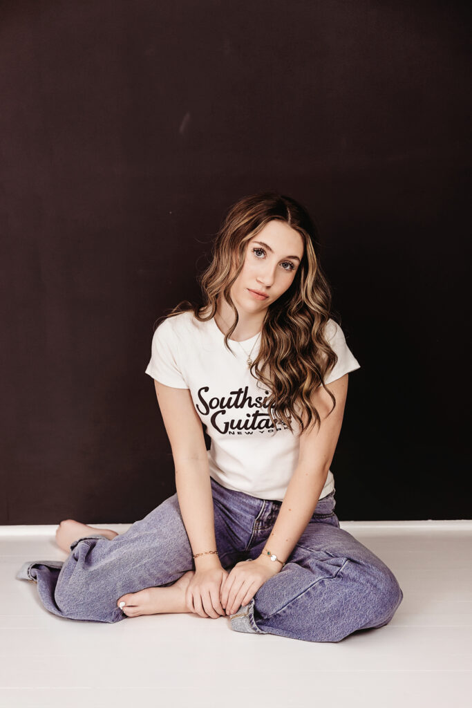 Teen girl seated cross-legged on the floor in front of a dark wall, wearing a 'Southside Guitars' tee with soft waves in her hair and a thoughtful expression.