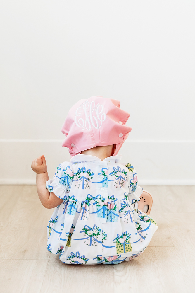 Back view of Elsie in floral romper, pink bonnet with monogram, sitting on studio floor at Reflection Images by Tracy Rowell in Florence SC