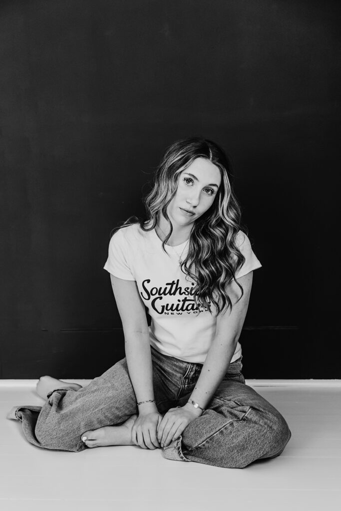 Black and white image of a teen girl sitting cross-legged in front of a black wall, hands resting in her lap, long hair curled.