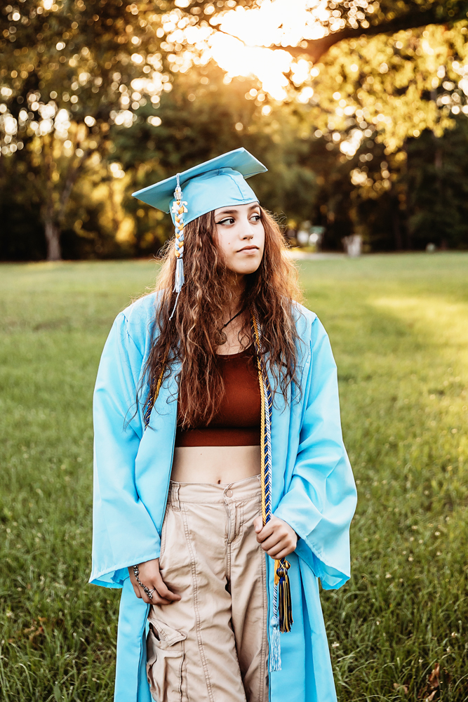 Eve standing in grassy field in blue cap and gown, rust top, khaki pants, golden sunset glow at Reflection Images by Tracy Rowell in Florence SC