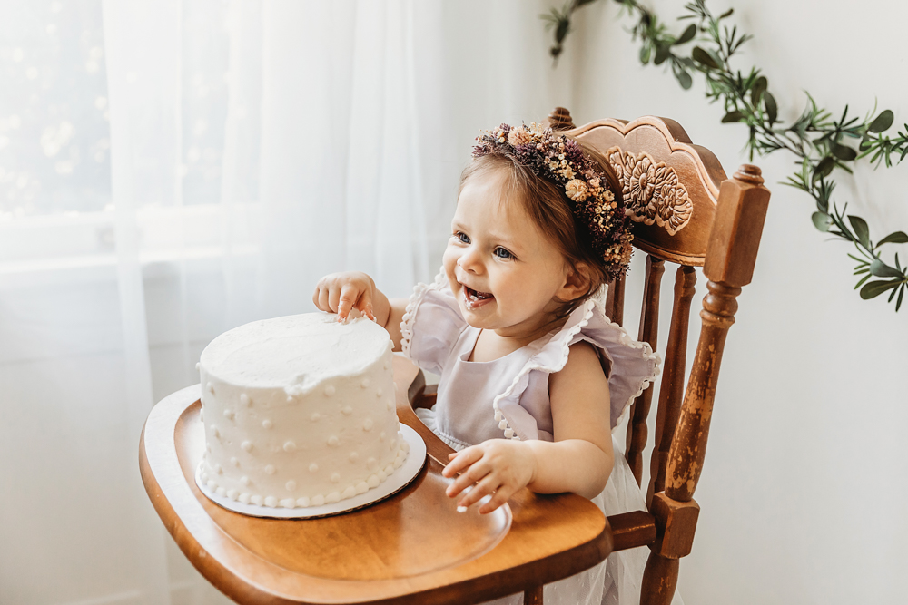 Smiling baby girl in a white dress looking at her cake while sitting in a wooden high chair during birthday celebration