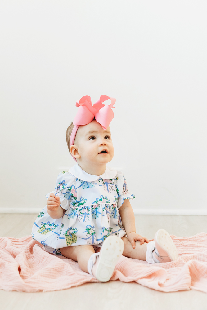 Elsie in floral romper with oversized pink bow, sitting on soft pink blanket, looking upward at Reflection Images by Tracy Rowell in Florence SC