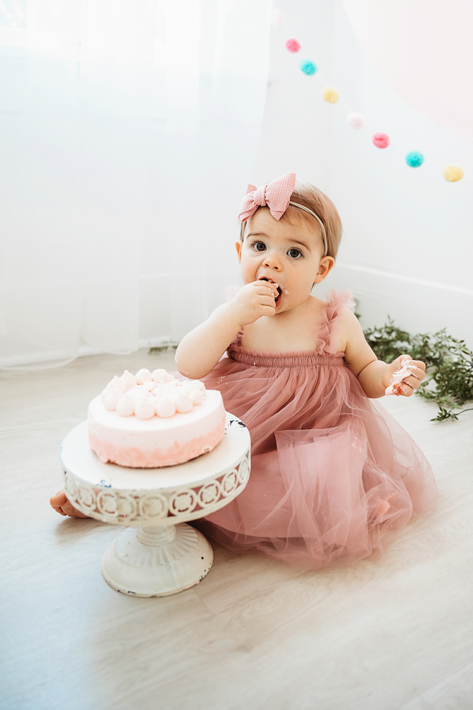 Close-up of Grace nibbling pink cake frosting with her fingers while wearing a pink tulle dress and birthday bow at Reflection Images by Tracy Rowell in Florence SC