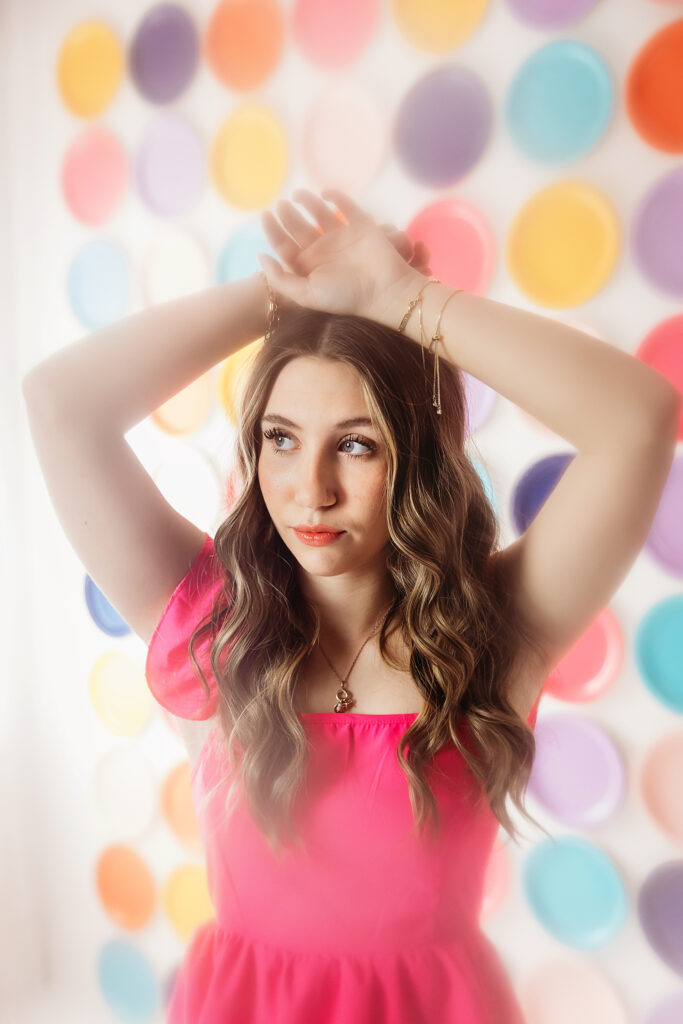 Teen girl with long waves wearing a pink dress standing in front of a dreamy wall filled with colorful circles, arms raised above her head.