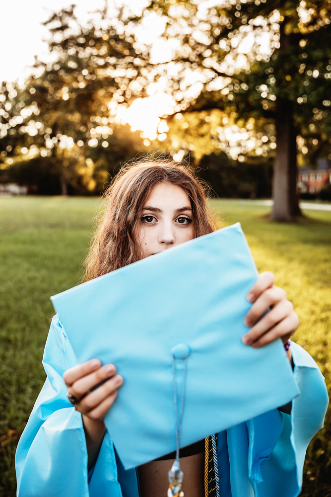 Eve holding blue graduation cap toward camera, eyes peeking above, golden sunlight and trees behind at Reflection Images by Tracy Rowell in Florence SC